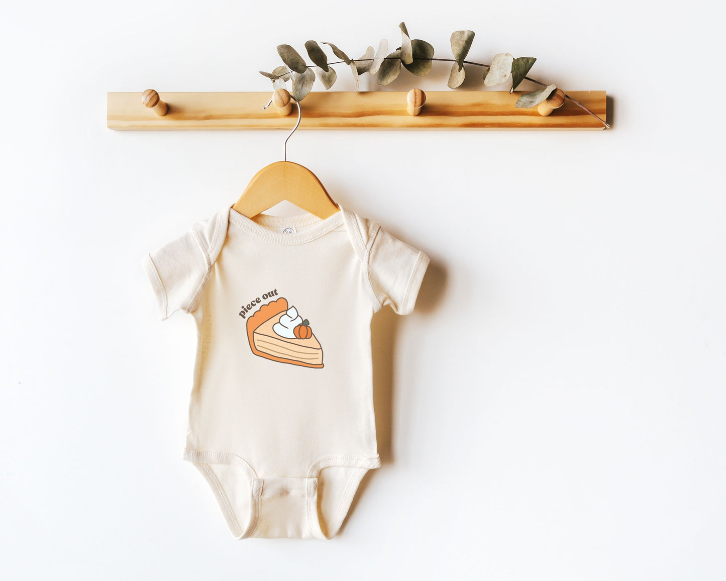 Short sleeve cotton baby onesie with a cute pumpkin pie drawing and the text "piece out" shown on a wooden hanger.