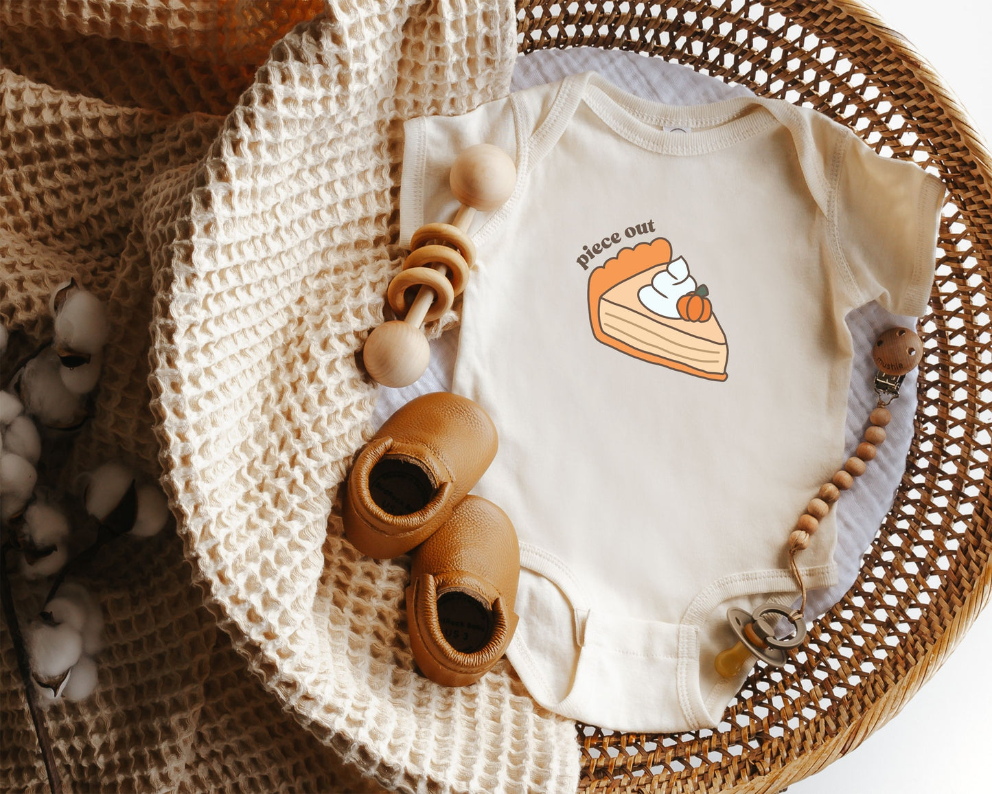 Short sleeve cotton baby onesie with a cute pumpkin pie drawing and the text "piece out" styled in a basket with a baby blanket, rattle and baby shoes.