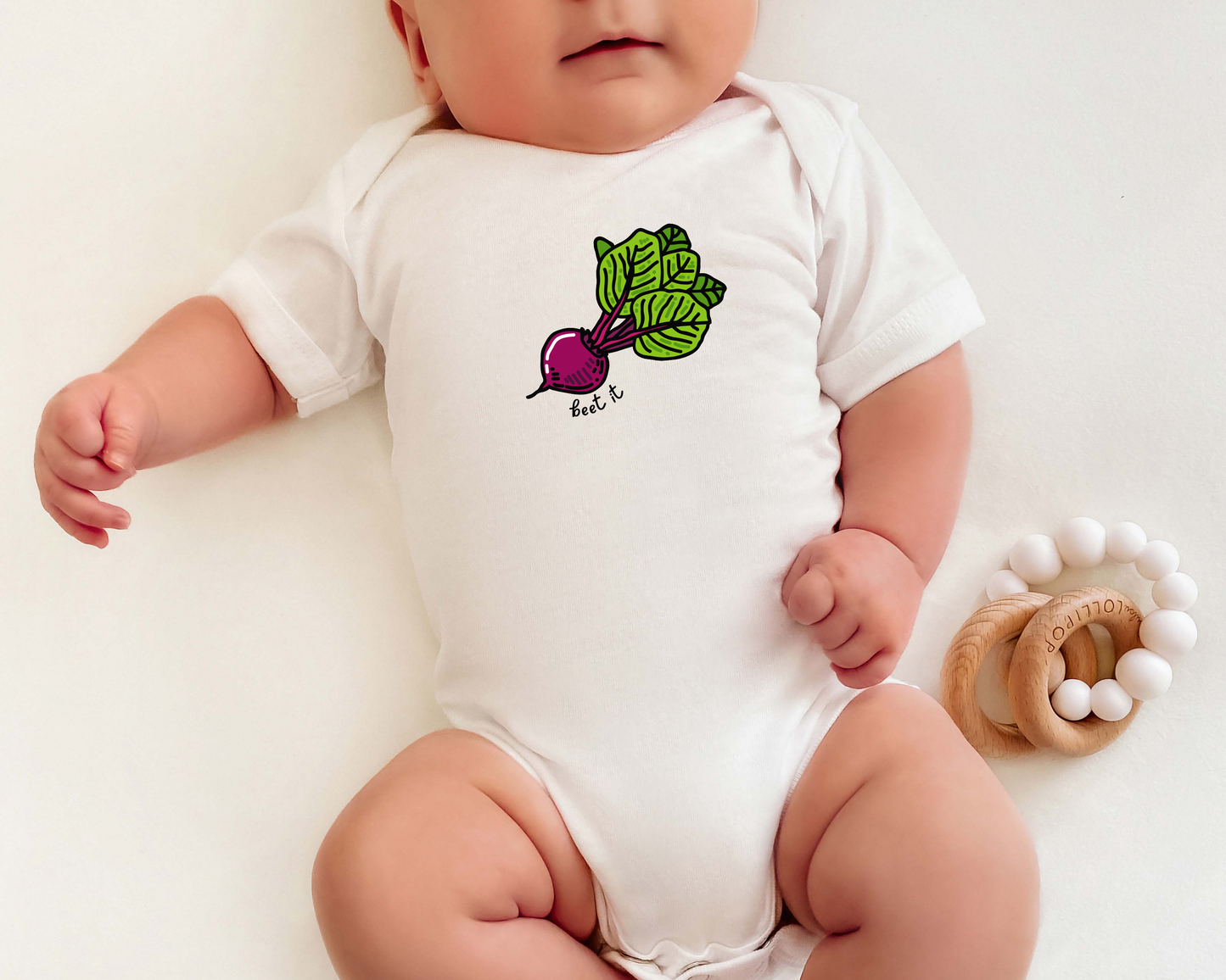 A baby wearing a white short-sleeve bodysuit with a colorful beet and leaf graphic on the chest, lying on a white background next to a wooden teether.