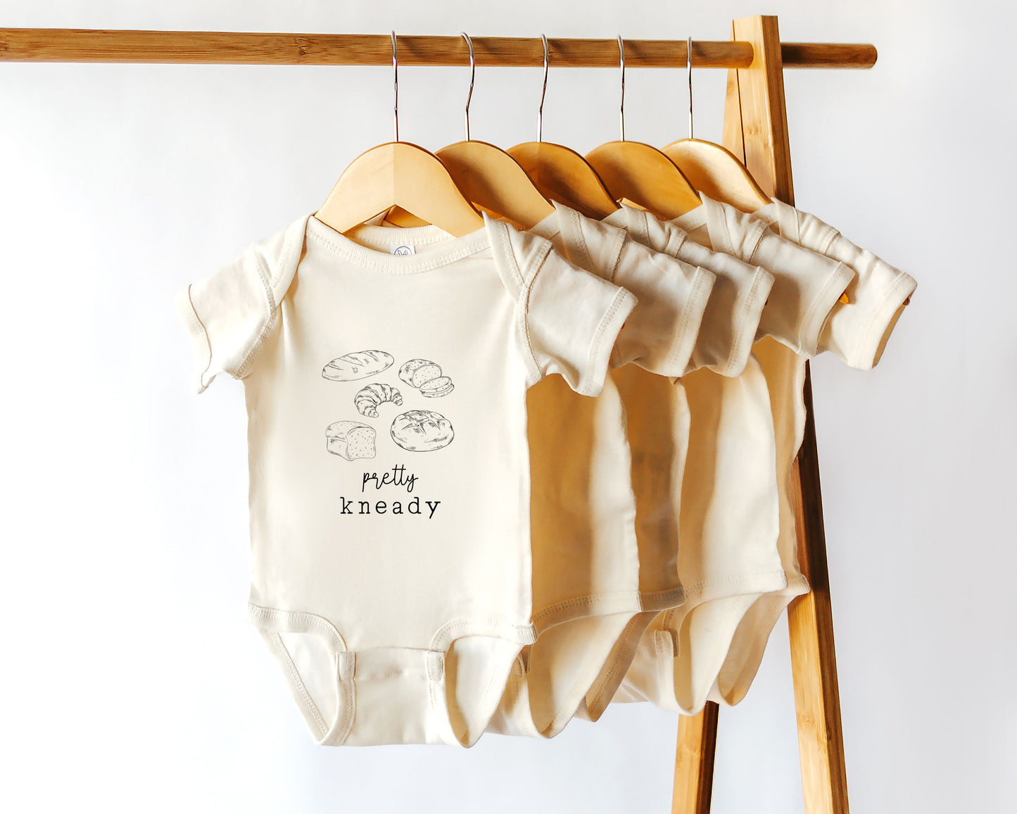 A short sleeve baby onesie with homemade bread illustrations and the phrase "pretty kneady"