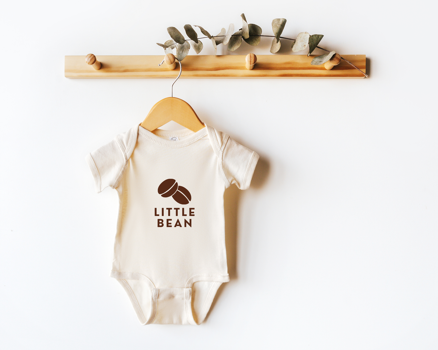 A short sleeve coffee themed baby onesie with a coffee bean graphic and the text little bean shown on a wooden hanger.