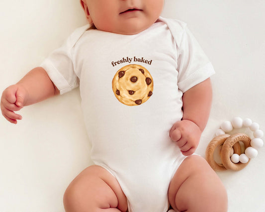 Short sleeve cotton baby onesie with a homemade chocolate chip cookie graphic and the text "freshly baked"