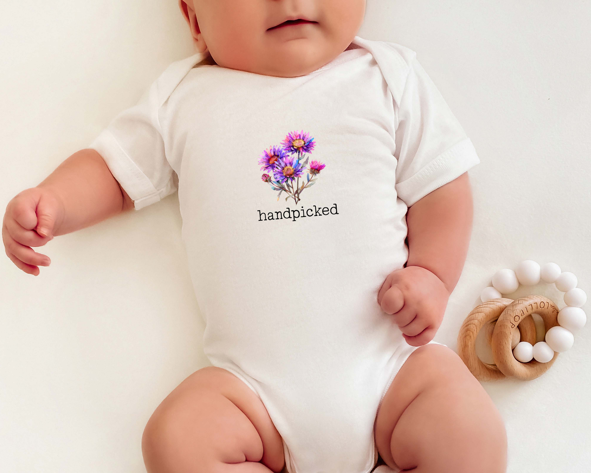 Short sleeve cotton baby onesie with graphic of aster flowers and the text "handpicked" in a typewriter style font.