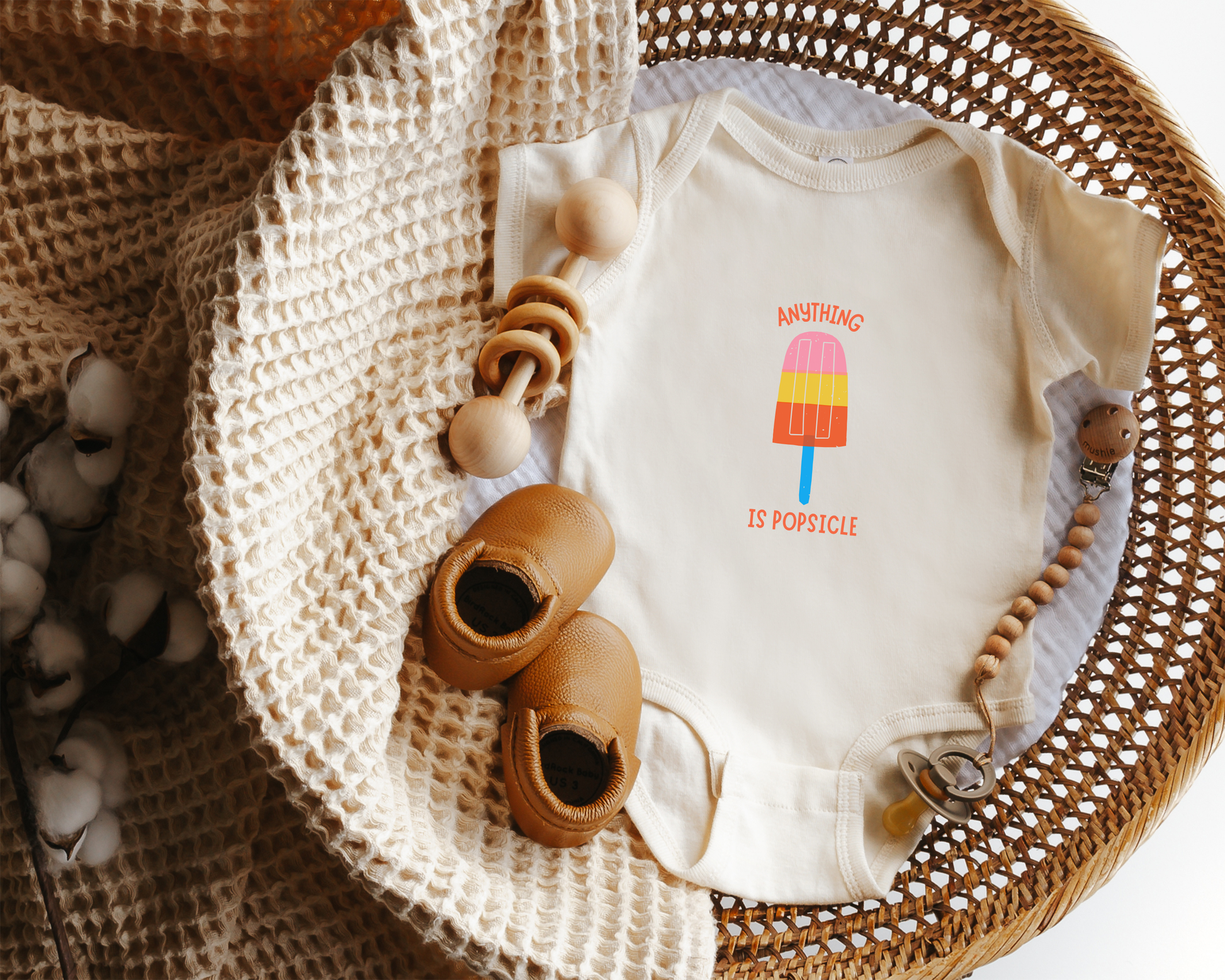 A short sleeve cotton baby onesie with a colorful popsicle graphic and the text "anything is popsicle" styled in a basket. 