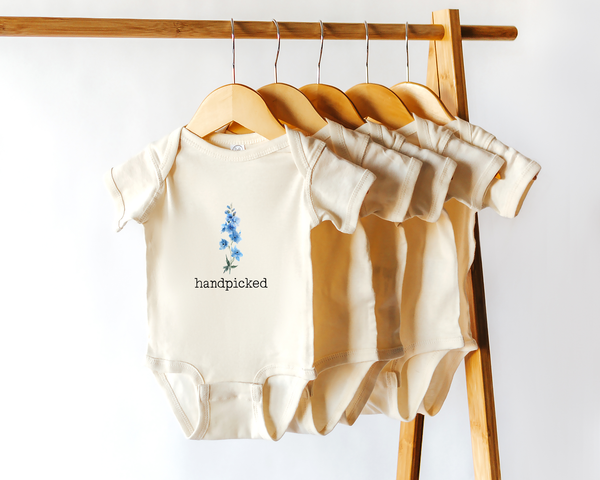 Short sleeve cotton baby onesie with graphic of blue lakespur flower and the text "handpicked"
