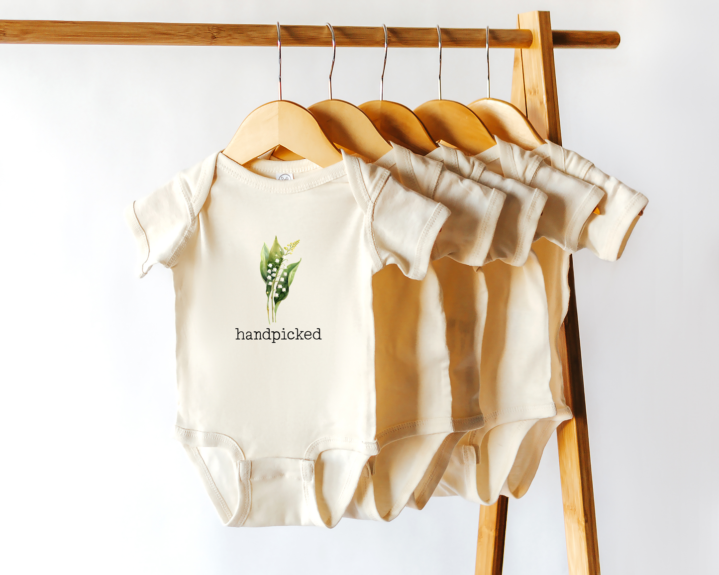 Short sleeve cotton baby onesie with a graphic of the May birth flower, Lily of the Valley, and the text "handpicked"