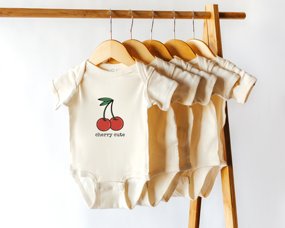 Baby onesie with cherry graphic and the text "cherry cute" on a wooden rack against a white background.