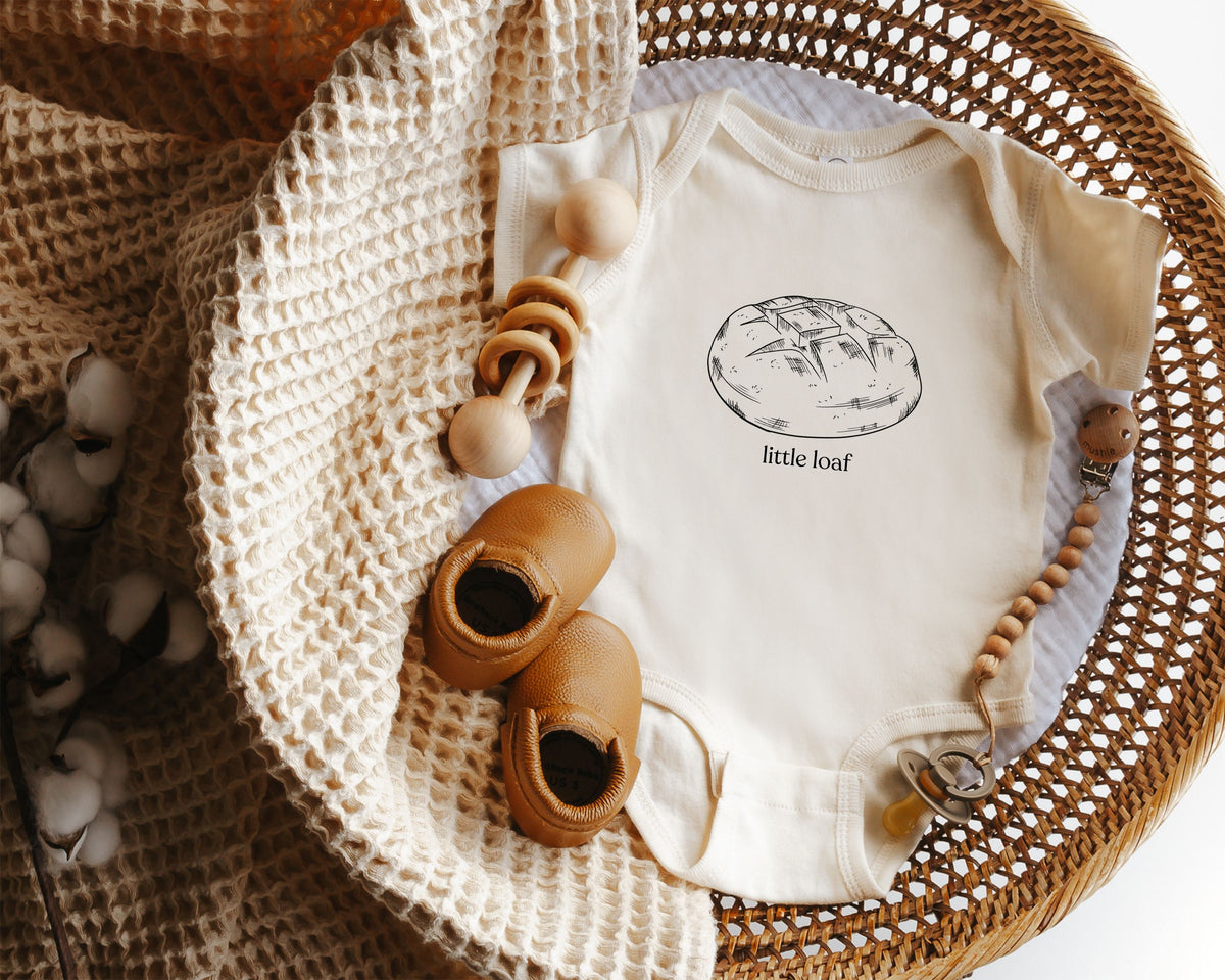 Baby onesie with a graphic of a sourdough loaf of bread and the text 'little loaf' displayed in a wicker basket.