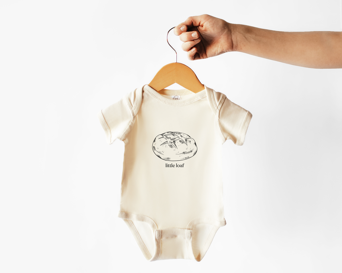 Baby onesie with a graphic of a sourdough bread loaf and the text 'little loaf' on a hanger against a white background