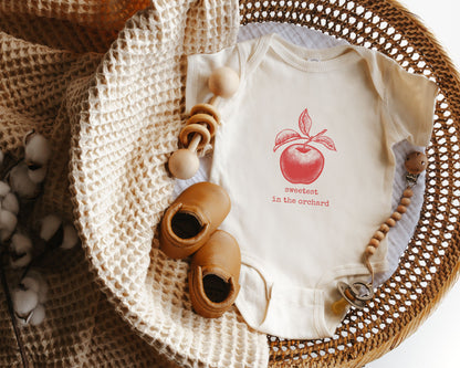 Short sleeve cotton onesie with a distressed style graphic of an apple in red with the text sweetest in the orchard in a typewriter style font.