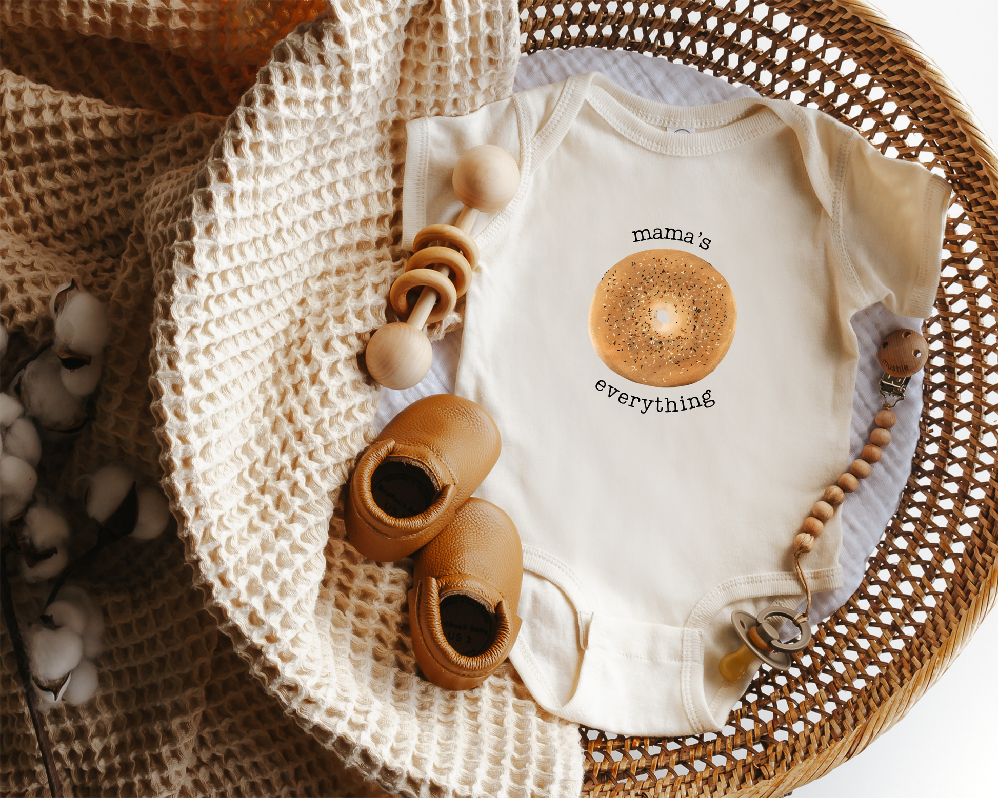 A short sleeve cotton baby onesie featuring an everything bagel graphic with the text "mama's everything" styled in a woven basket.