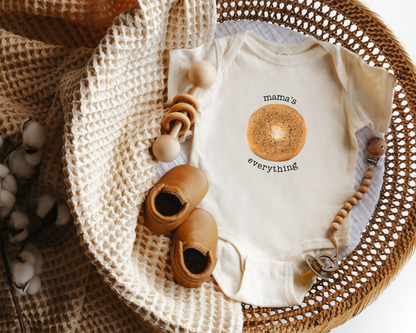 A short sleeve cotton baby onesie featuring an everything bagel graphic with the text "mama's everything" styled in a woven basket.