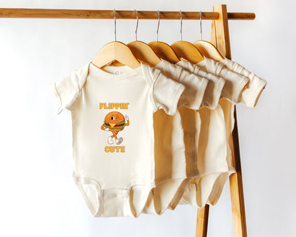 A cotton baby onesie with a retro style burger graphic and the text "flippin' cute" displayed on a wooden hanger. 