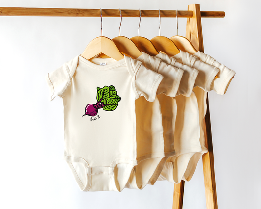 A set of cream-colored short-sleeve baby bodysuits hanging on wooden hangers from a wooden clothing rack. The front bodysuit features a graphic of a beet with green leaves and the text 'beet it'.
