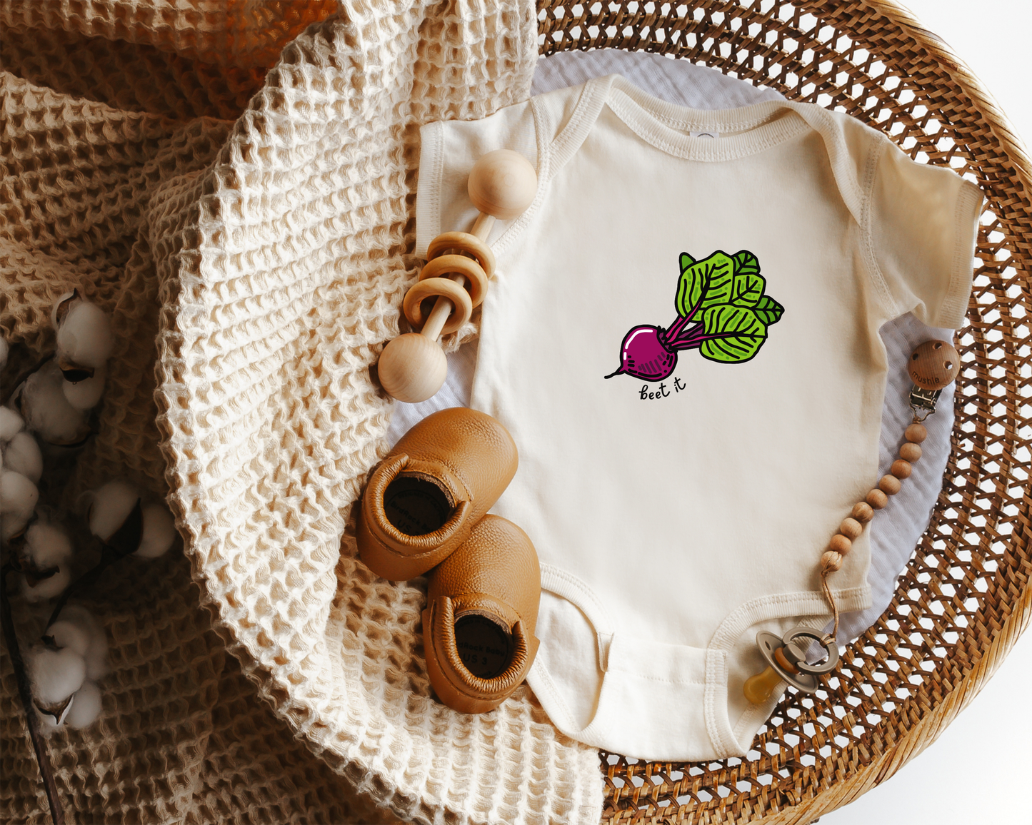 An off-white baby bodysuit with a beet graphic on the chest, styled in a wicker basket with wooden toys, a knit blanket, and baby shoes.