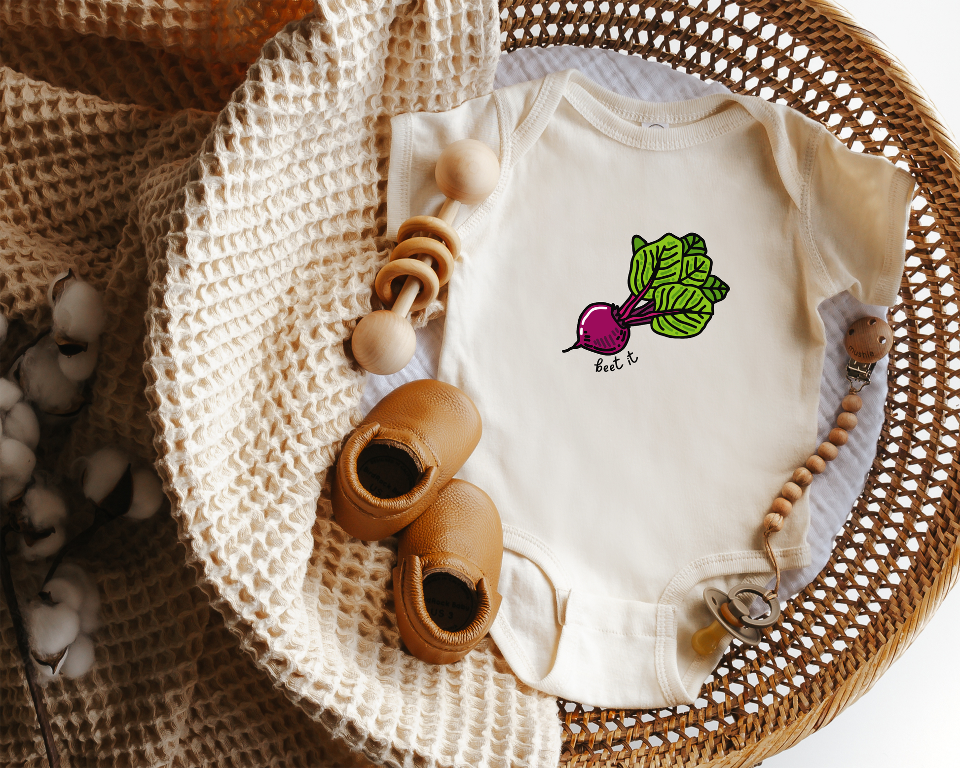 An off-white baby bodysuit with a beet graphic on the chest, styled in a wicker basket with wooden toys, a knit blanket, and baby shoes.