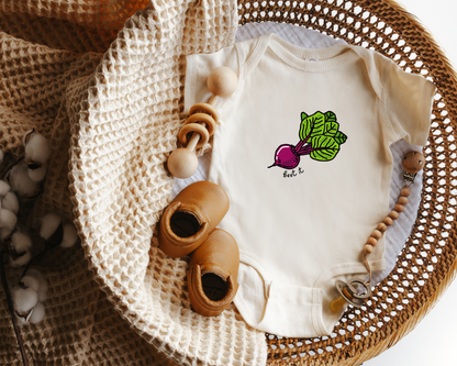 An off-white baby bodysuit with a beet graphic on the chest, styled in a wicker basket with wooden toys, a knit blanket, and baby shoes.