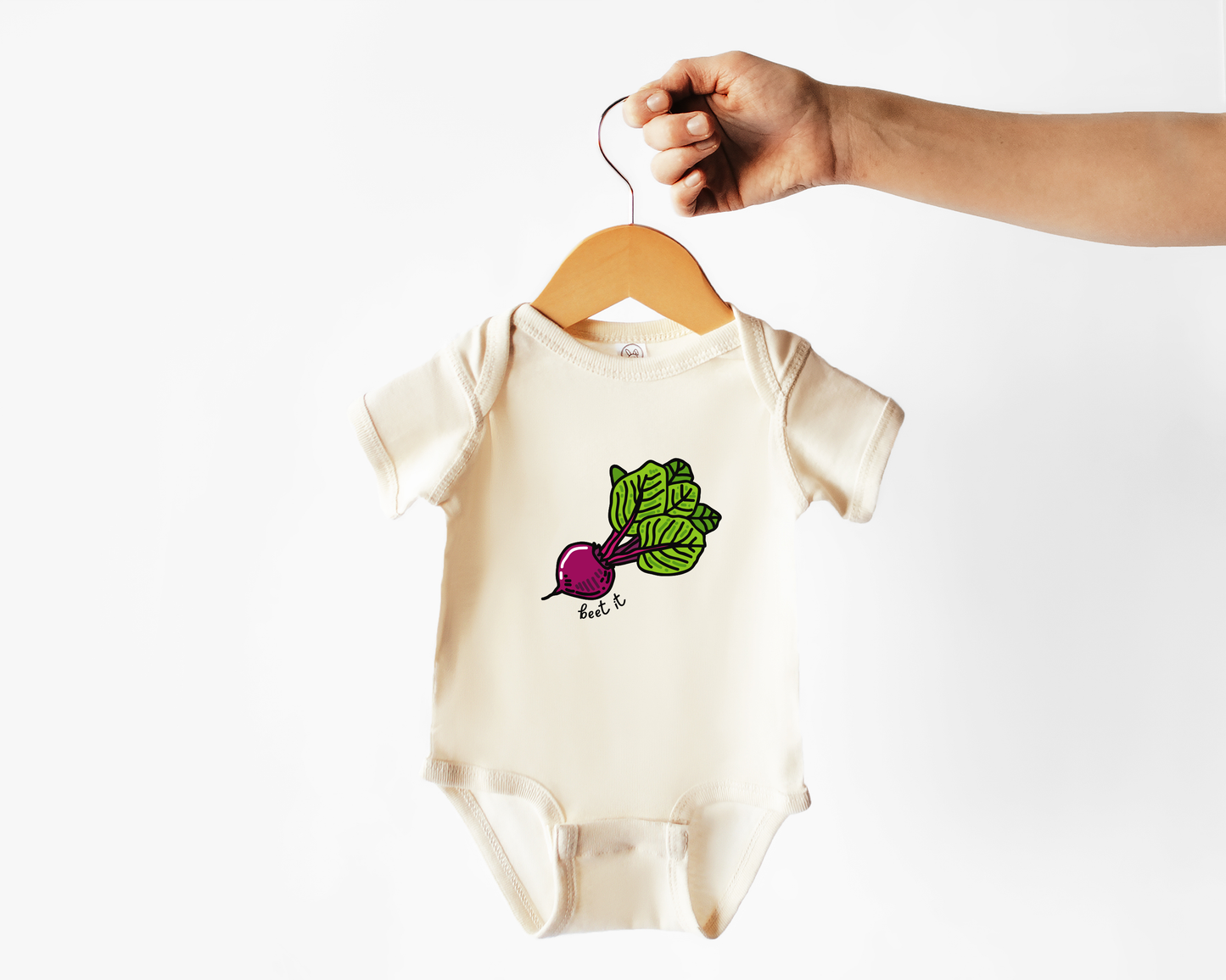 A cream-colored short-sleeve baby bodysuit with a beet and leaf graphic on the chest, hanging from a wooden hanger held by a person's hand against a plain white background.