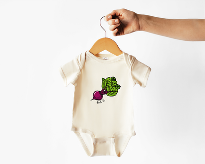 A cream-colored short-sleeve baby bodysuit with a beet and leaf graphic on the chest, hanging from a wooden hanger held by a person's hand against a plain white background.