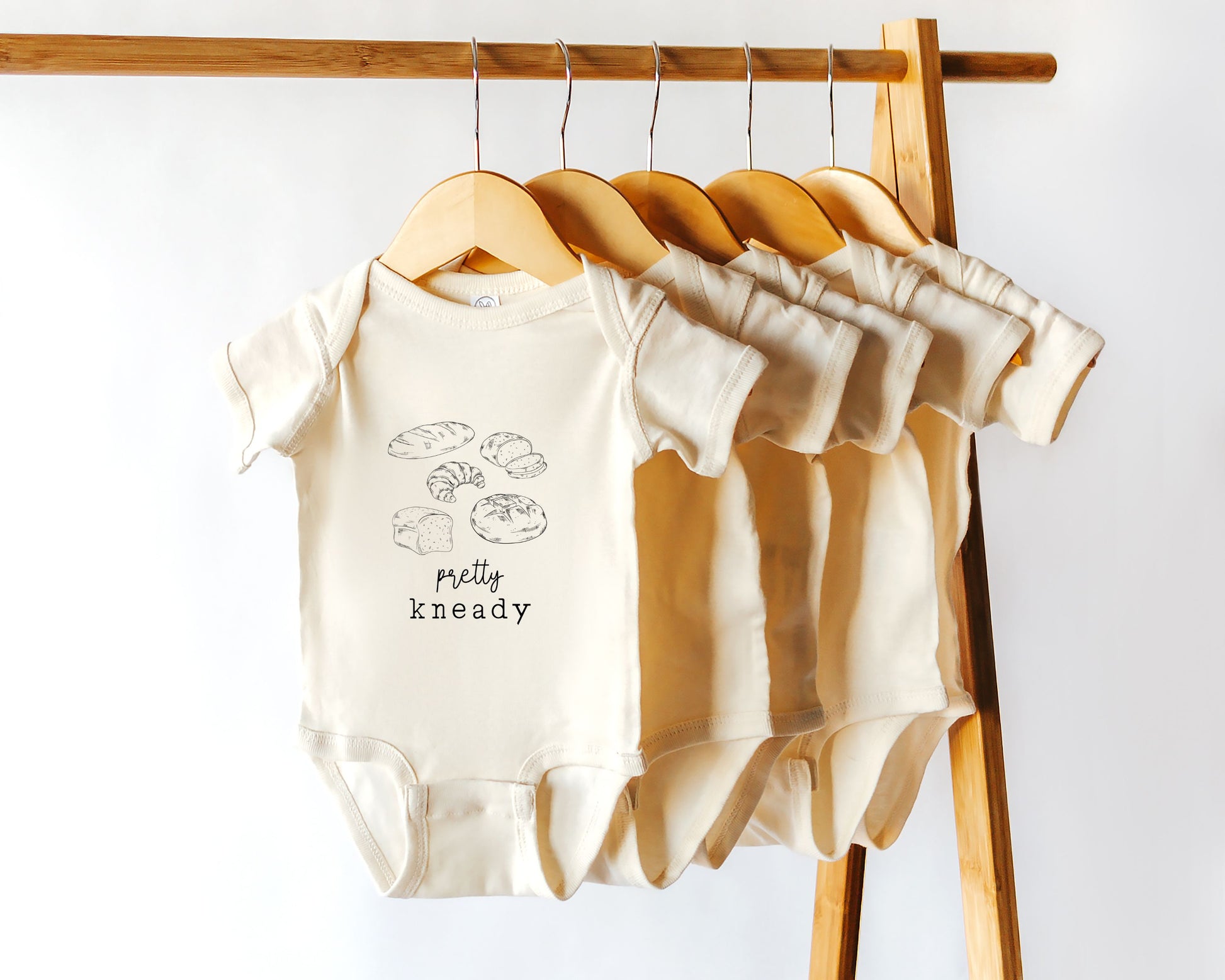 A short sleeve baby onesie with homemade bread illustrations and the phrase "pretty kneady"