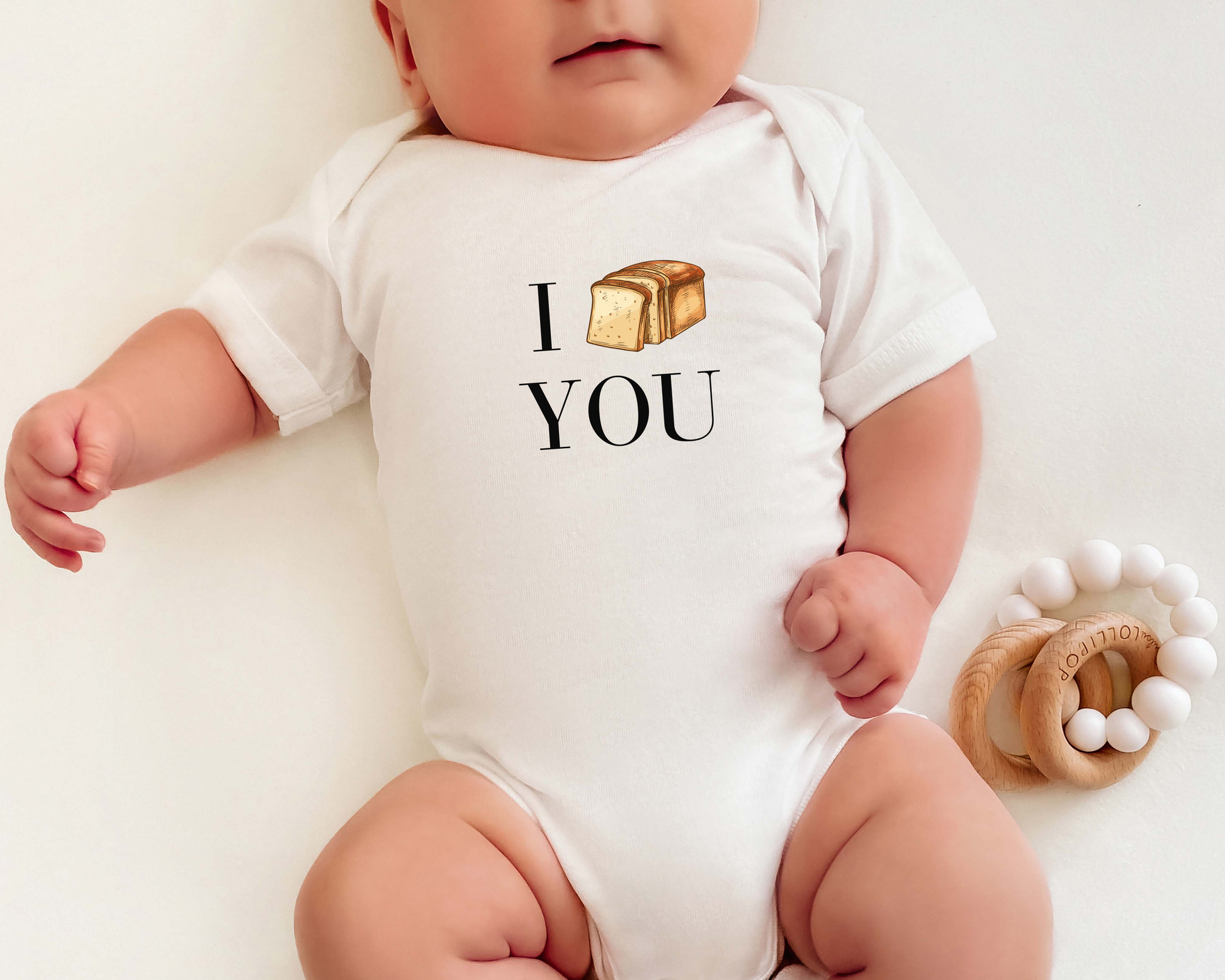 A baby wearing a white short-sleeve bodysuit with a graphic that reads 'I LOAF YOU' in black text. The baby is lying on a white surface next to a wooden teething ring with white beads.