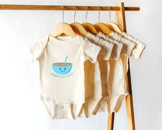 A cotton baby onesie with a cereal bowl graphic and the text "cereal-sly cute". Displayed on a wooden hanger.