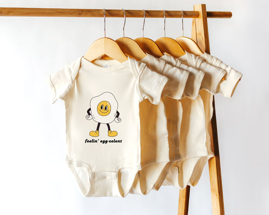 A short sleeve cotton baby onesie with a retro style egg character graphic and the text "feelin' egg-celent" styled on a wooden rack among other similar bodysuits.