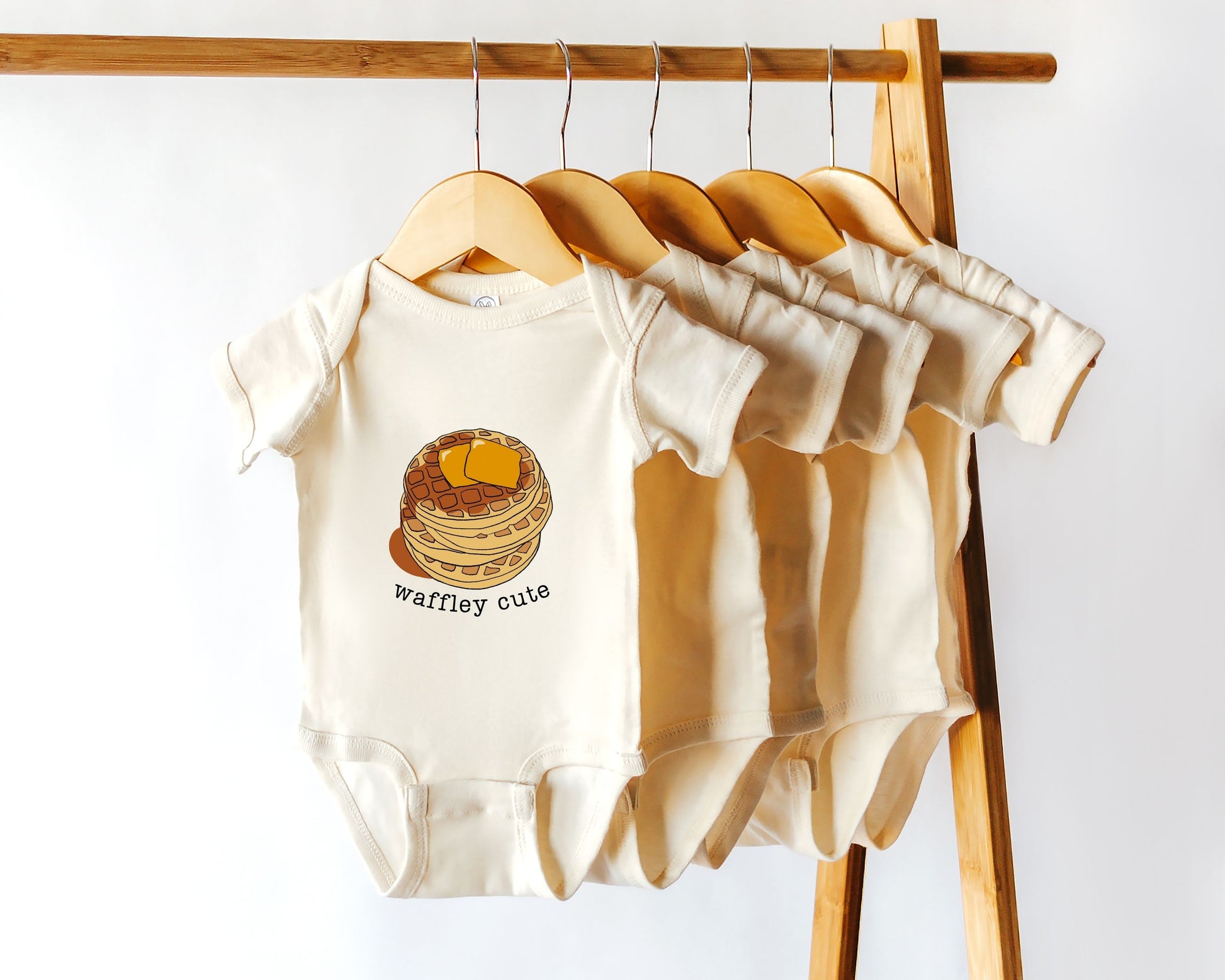 A cotton baby onesie with the text 'Waffley Cute' printed on the front with a stack of waffles graphic displayed among other bodysuits on a wooden hanger.