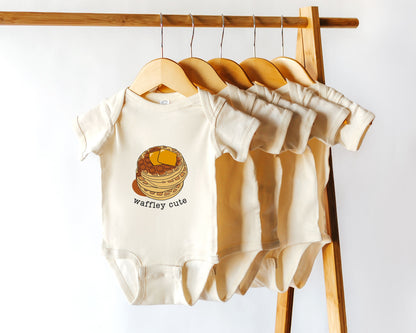 A cotton baby onesie with the text 'Waffley Cute' printed on the front with a stack of waffles graphic displayed among other bodysuits on a wooden hanger.