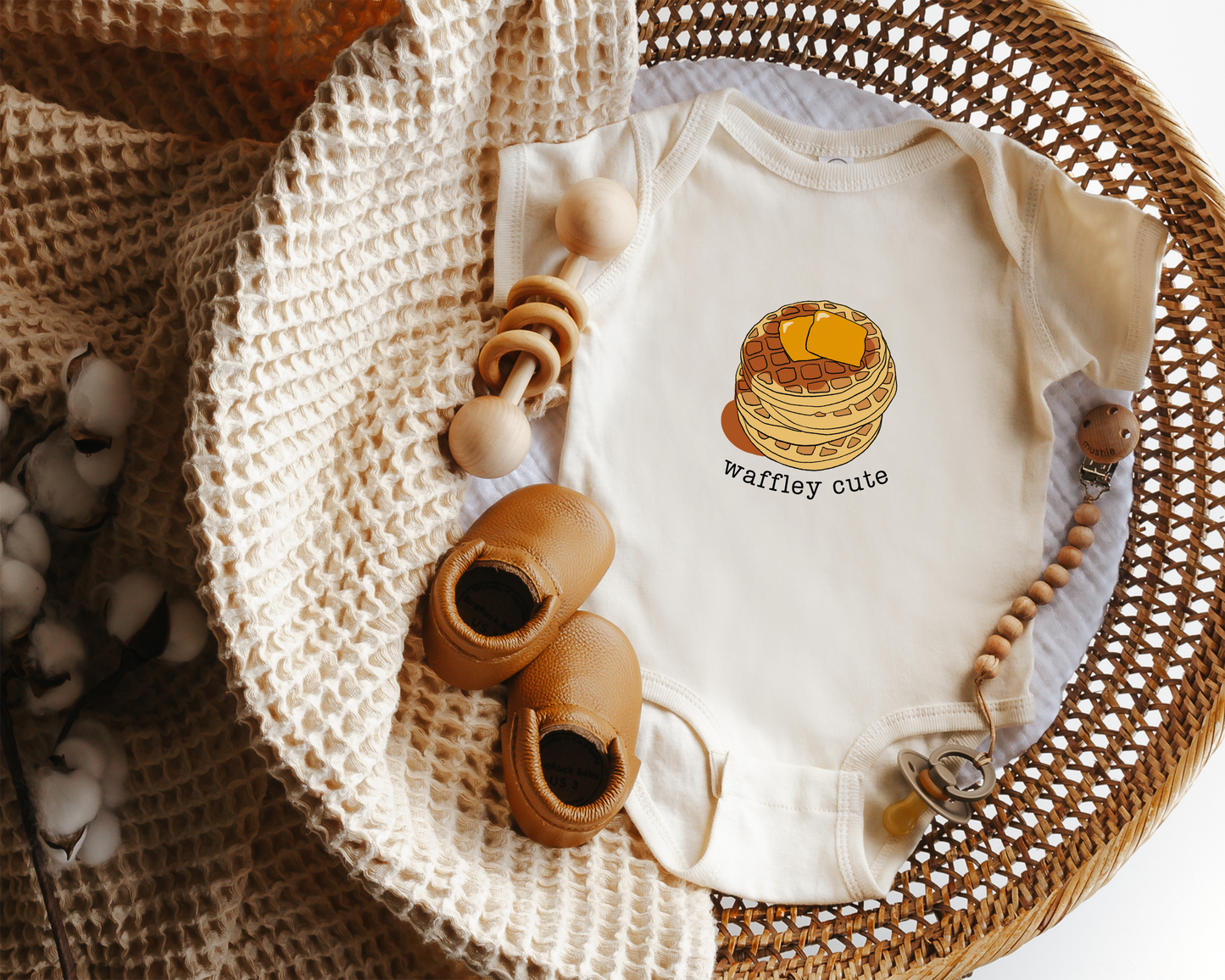 A baby onesie laid flat in a woven basket with styled props including a knit blanket, wooden toy and baby shoes. The onesie is cream with a stack-of-waffles graphic and the words 'waffley cute'.