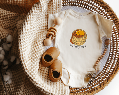 A baby onesie laid flat in a woven basket with styled props including a knit blanket, wooden toy and baby shoes. The onesie is cream with a stack-of-waffles graphic and the words 'waffley cute'.