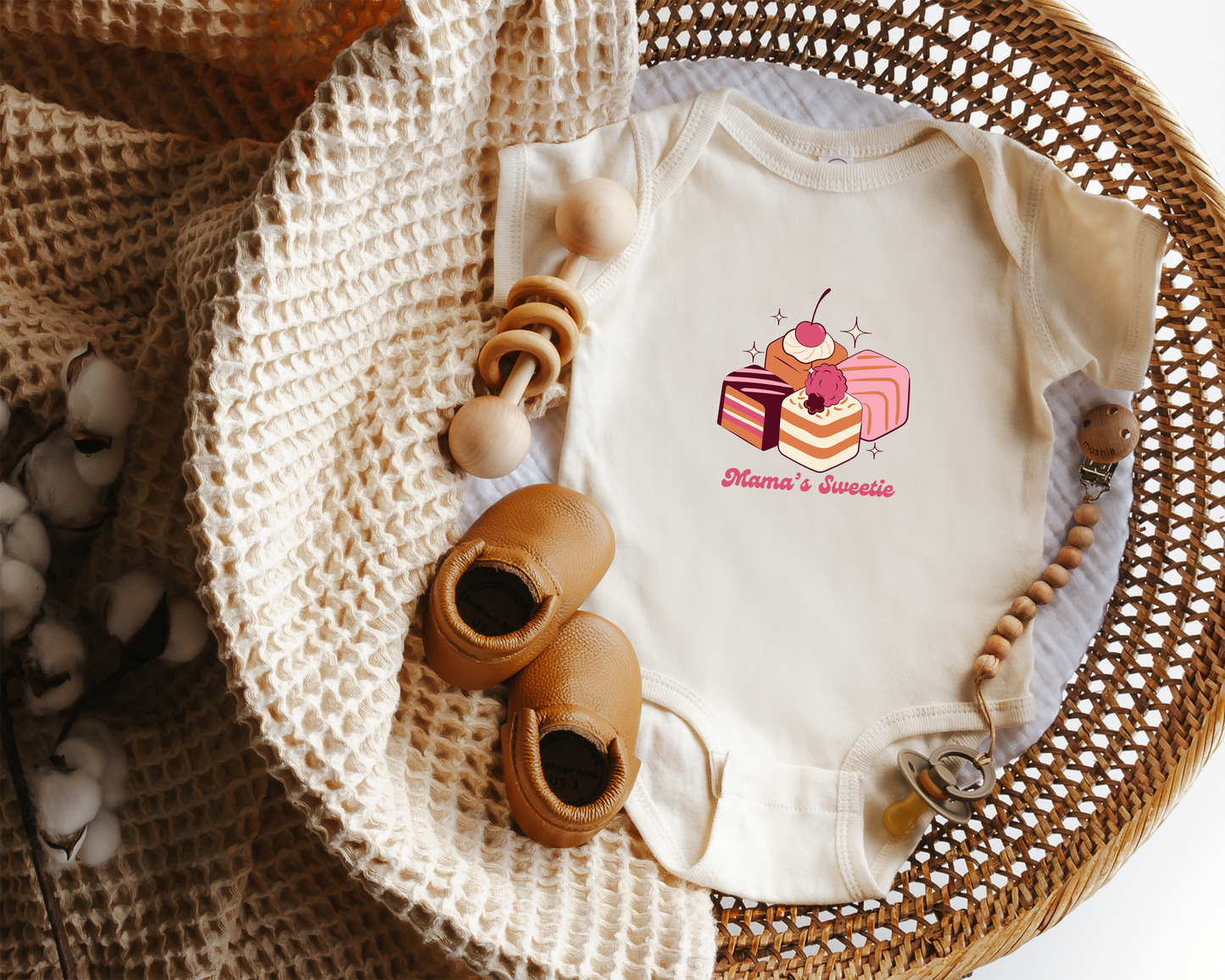Baby onesie with cake graphics and 'Mama's Sweetie' text displayed in a wicker basket. 