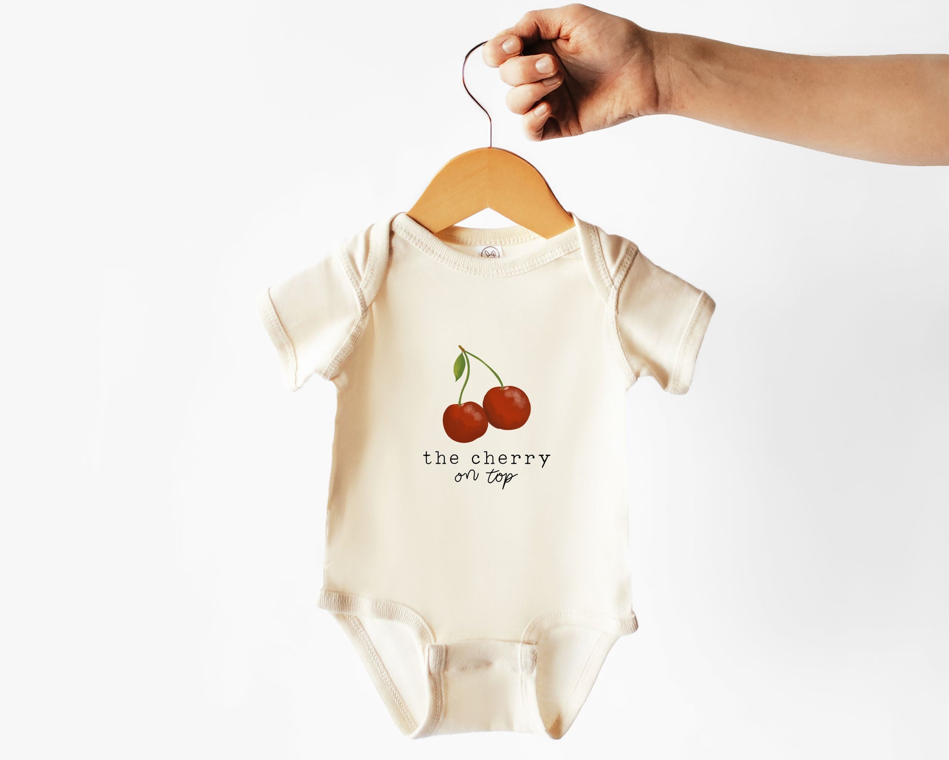 Baby onesie with cherry graphic and the text "the cherry on top" shown on a hanger against a white background
