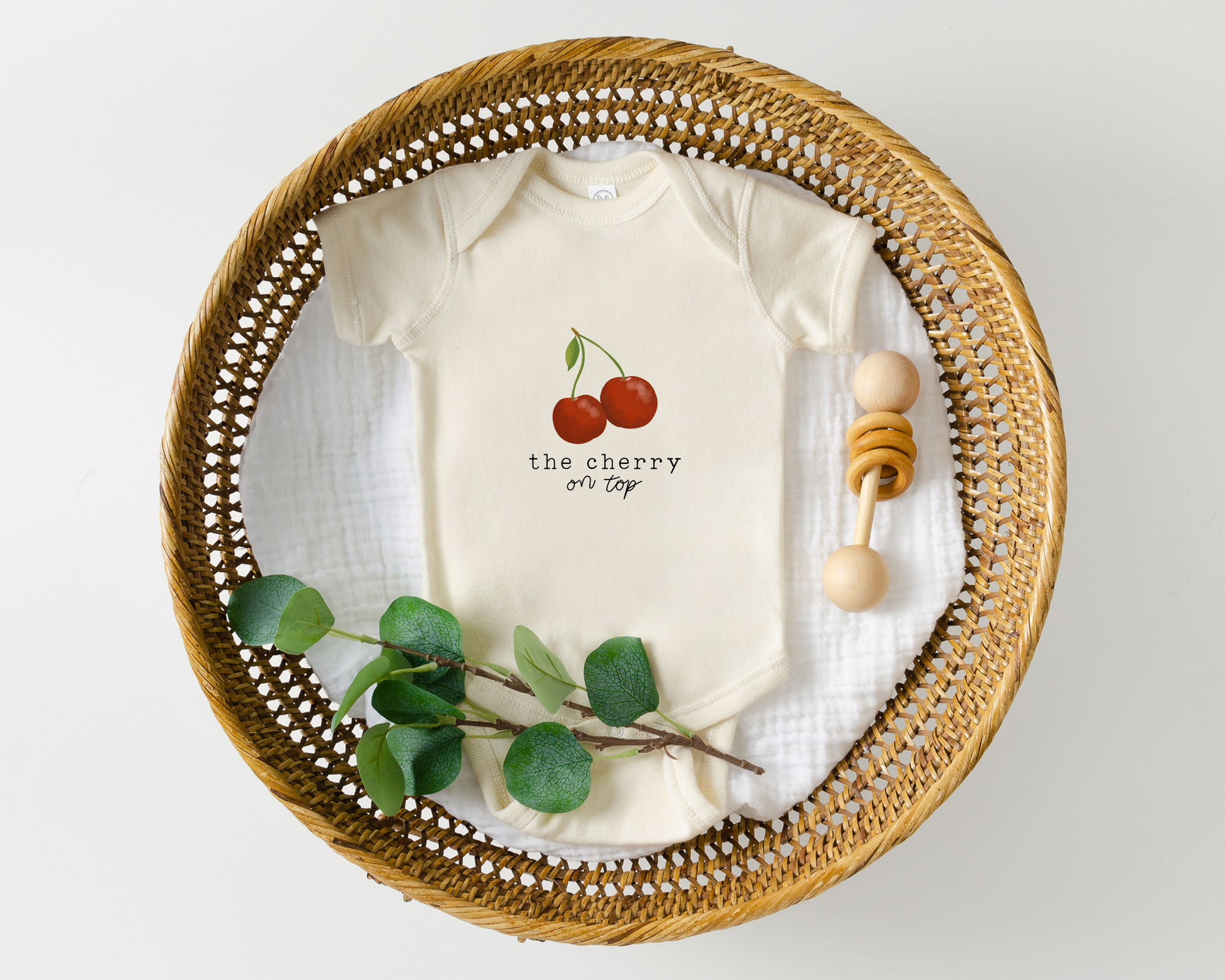 Baby onesie with cherry graphic and the text "the cherry on top" styled in a basket with wooden baby toy.