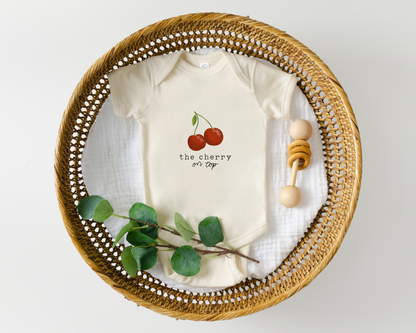 Baby onesie with cherry graphic and the text "the cherry on top" styled in a basket with wooden baby toy.