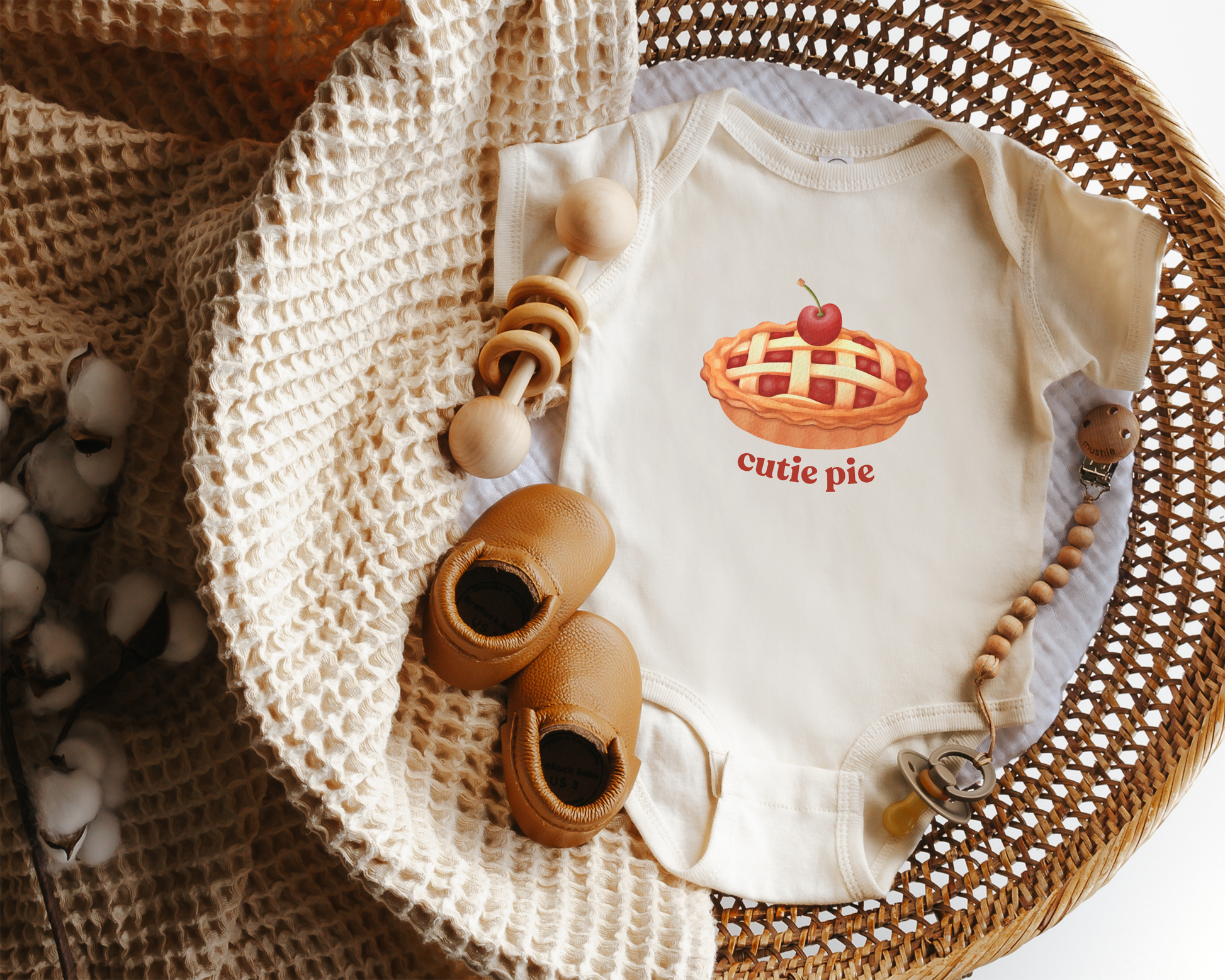 A cream-colored baby bodysuit featuring a 'cutie pie' graphic, styled in a flat-lay arrangement inside a wicker basket with a knit blanket, wooden toy and baby shoes.