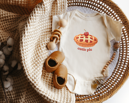 A cream-colored baby bodysuit featuring a 'cutie pie' graphic, styled in a flat-lay arrangement inside a wicker basket with a knit blanket, wooden toy and baby shoes.