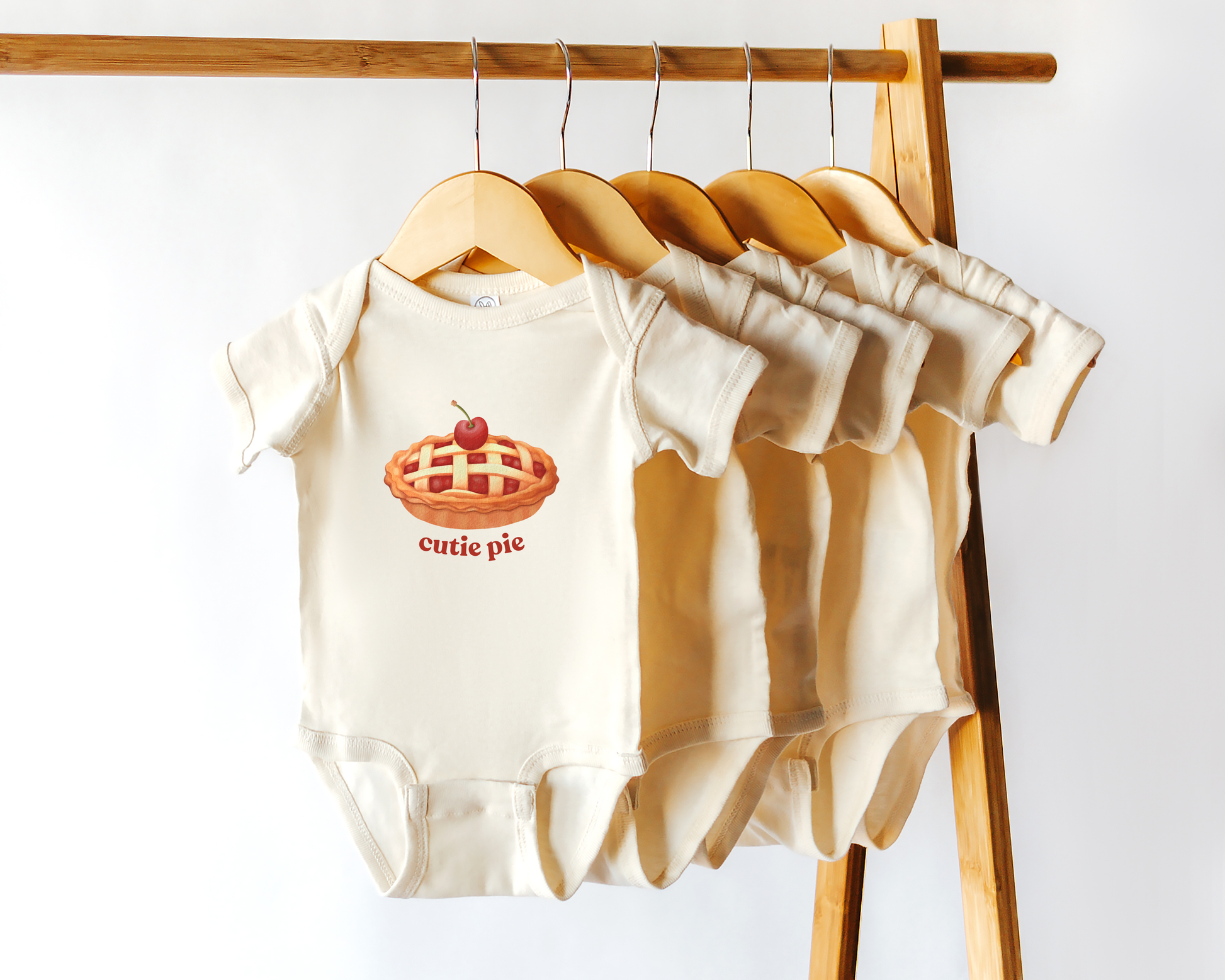 A set of cream-colored baby bodysuits hanging on wooden hangers from a wooden clothing rack. The front bodysuit features a graphic of a cherry pie with the text 'cutie pie' printed in red.