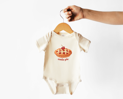 A cream-colored baby short-sleeve bodysuit hanging from a wooden hanger held by a hand, featuring a graphic of a cherry pie with the text 'cutie pie' underneath.