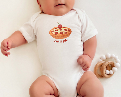 A baby wearing a white short-sleeve bodysuit with a graphic of a cherry pie and the text 'cutie pie' on the chest, lying on a white background next to a wooden teething ring.