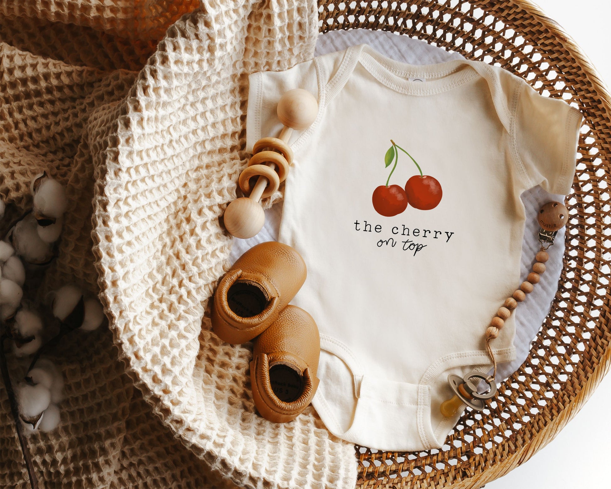 Baby onesie with cherry graphic and the text "the cherry on top" styled in a wicker basket.