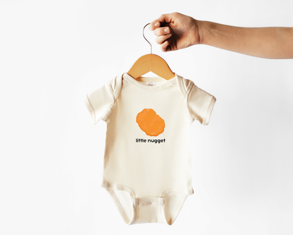 A cream-colored baby bodysuit with a chicken nugget graphic and the text 'little nugget' hanging from a wooden hanger held by a hand against a plain white background.