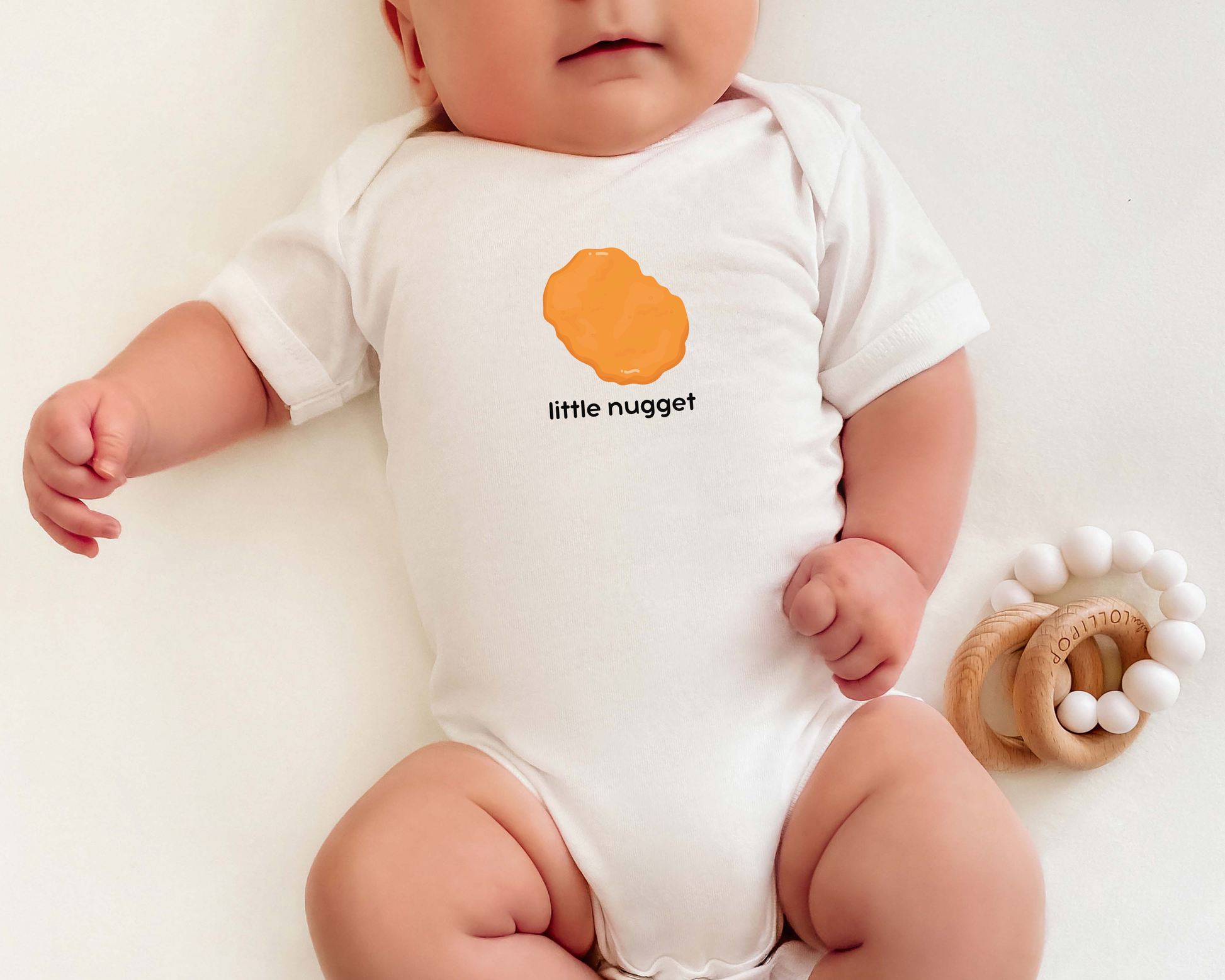 A baby wearing a white short-sleeve bodysuit with a chicken nugget graphic and the text 'little nugget' on it.