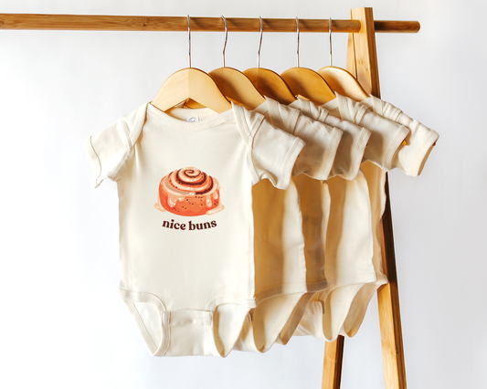 A short sleeve cotton baby onesie with a cinnamon bun graphic and the text "nice buns" displayed on a wooden hanger.