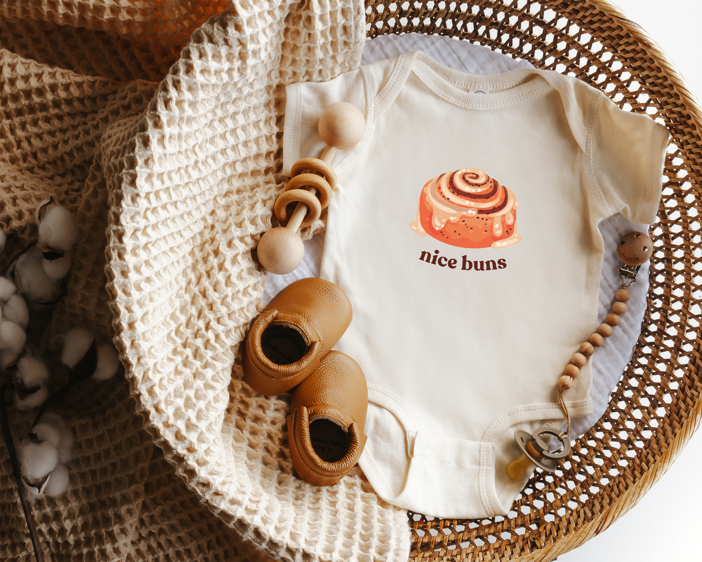 A short sleeve baby onesie with a cinnamon bun graphic and the text "nice buns" styled in a wicker basket. 