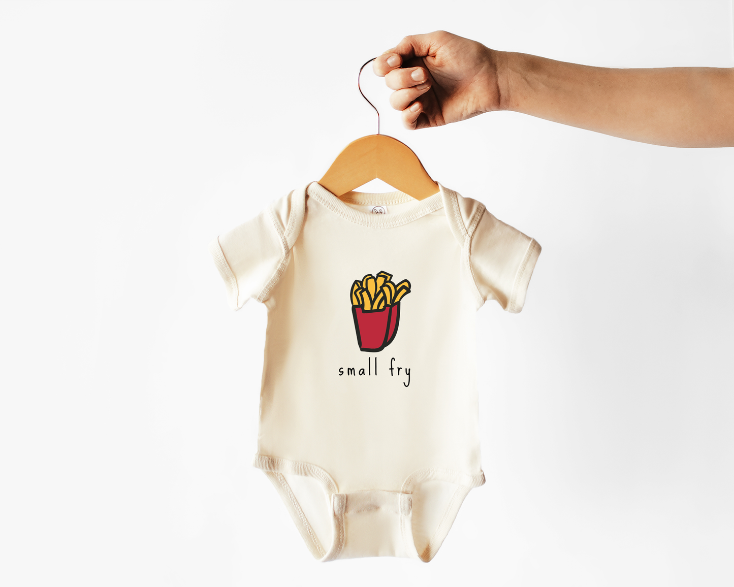 A baby short-sleeve bodysuit in cream, hanging on a wooden hanger held by a hand. The bodysuit features a graphic of french fries in a red bucket with the text 'small fry' underneath.