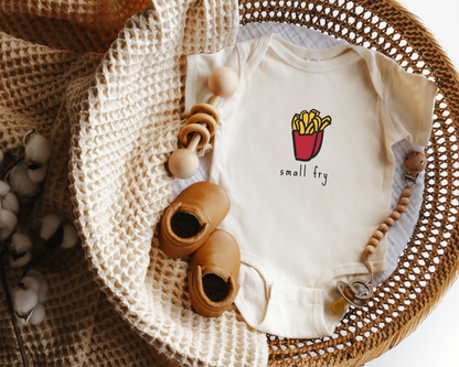 A cream-colored baby bodysuit with a 'small fry' french fries graphic, styled in a a woven basket with baby shoes and a knit blanket.