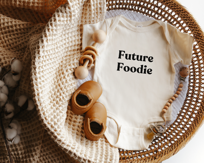 A cream-colored baby bodysuit with the words 'Future Foodie' printed in black on the chest, styled in a woven basket.