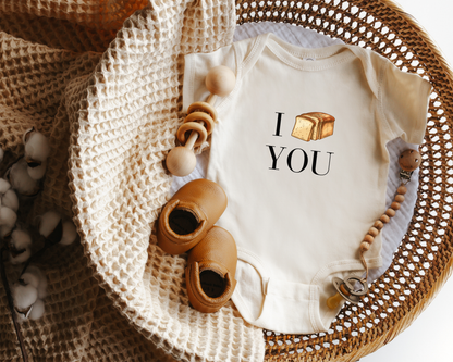 A baby bodysuit with the text 'I [bread slice graphic] YOU' in black, styled in a woven basket. 