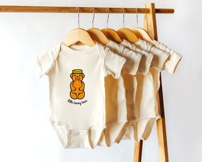 A natural baby bodysuit with a graphic of a honey bear jar on the front, displayed on a wooden hanger among other similar hangers.